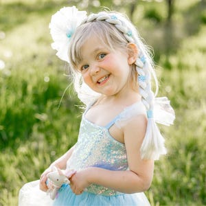 May include: A light blue sequined dress with a tulle skirt and a white braided hair extension accessory featuring small blue floral accents and a large white tulle hair flower, paired with a small plush unicorn toy held in the hands.
