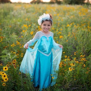May include: A bright turquoise dress featuring a sequined bodice, sheer long sleeves, and a flowing skirt with a matching sheer cape. The outfit is accessorized with a white snowflake-shaped hair accessory and a silver pendant necklace.