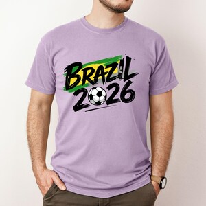 Op de afbeelding: Een lichtpaars t-shirt met de tekst "BRAZIL 2026" in zwart, met een voetbalafbeelding. De tekst staat boven een groen en geel penseelstrekenontwerp, dat de Braziliaanse vlag vertegenwoordigt.