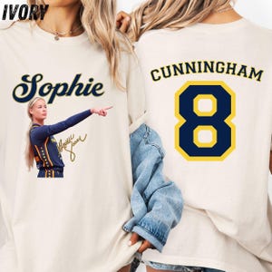 May include: An ivory t-shirt featuring a graphic of a basketball player pointing, the name Sophie, and a signature on the front. The back displays the name Cunningham in dark blue letters above a large dark blue and yellow number 8 graphic.