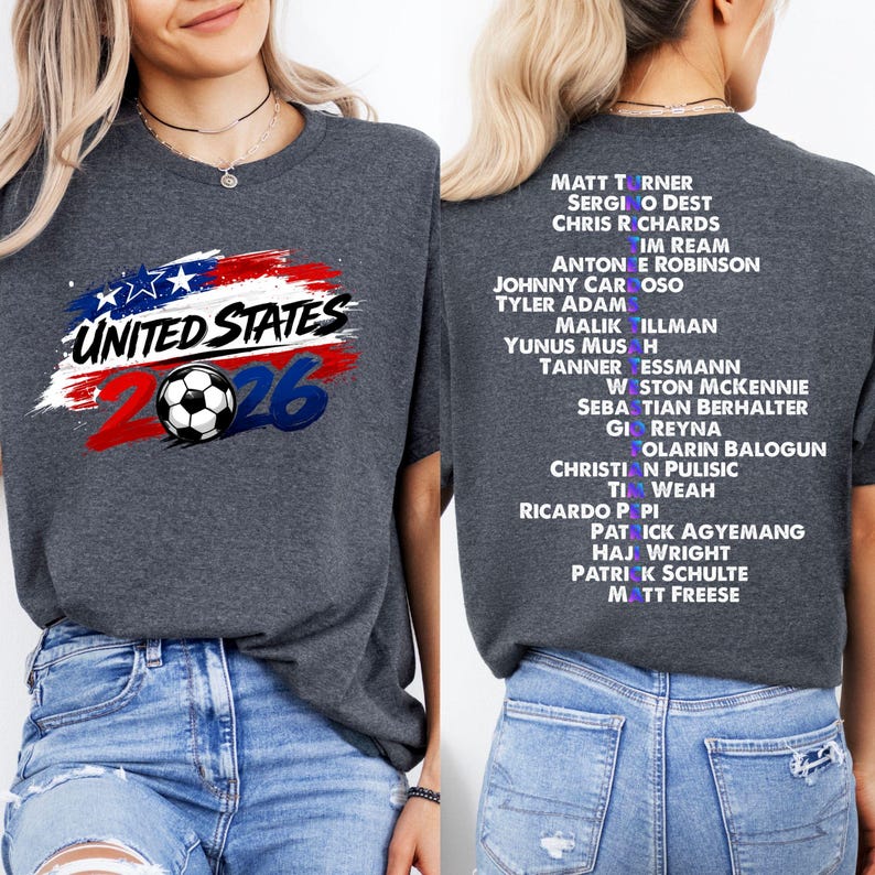 May include: A dark grey t-shirt with a graphic for the United States 2026 soccer event. The front displays "United States" with a soccer ball and "26". The back has a list of player names.
