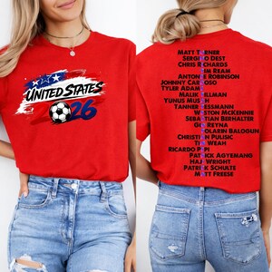 May include: Red t-shirt with a soccer ball graphic and the text "UNITED STATES 2026". The back of the shirt features a list of names. The shirt is paired with blue jeans.