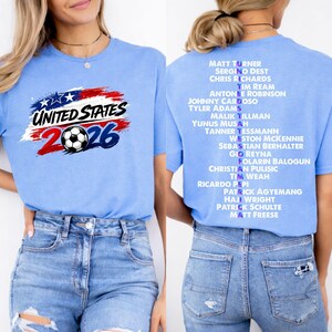 May include: Pale blue t-shirt featuring the text "UNITED STATES 2026" and a football graphic. The back of the shirt lists soccer player names. The design incorporates the colours of the American flag.