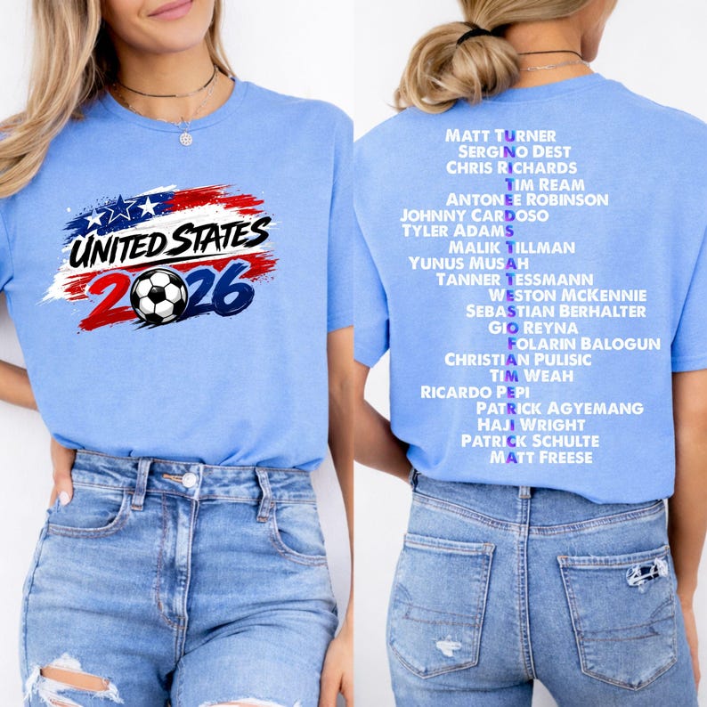 May include: Pale blue t-shirt featuring the text "UNITED STATES 2026" and a football graphic. The back of the shirt lists soccer player names. The design incorporates the colours of the American flag.