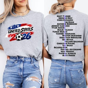 May include: A grey t-shirt with the text "UNITED STATES 2026" and a football graphic. The back of the shirt lists soccer player names. The design uses red, white, and blue colours.