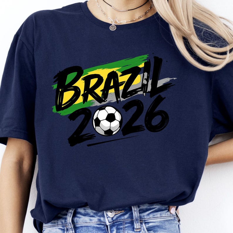 Op de afbeelding: Marineblauw T-shirt met het woord "BRAZIL" in zwart, met een geel, groen en grijs penseelstreken ontwerp. Onder het woord "BRAZIL" staat het nummer "2026" en een voetbalafbeelding.