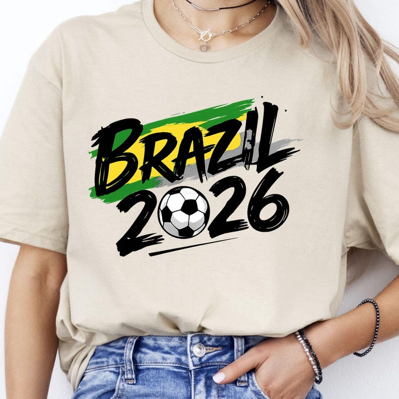 Op de afbeelding: Beige T-shirt met het woord "BRAZIL" in zwart, met een ontwerp van groene, gele en grijze strepen. Onder het woord "BRAZIL" staat het nummer "2026" en een voetbal.