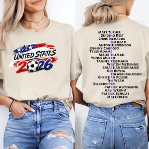 May include: Beige t-shirt with the text "UNITED STATES 2026" and a soccer ball graphic. The back of the shirt lists soccer player names. The shirt is made of a soft material.