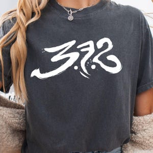 May include: Dark gray t-shirt with the number "372" in white, stylized brushstroke font. The shirt has a crew neck and short sleeves. A silver necklace is visible at the neckline.