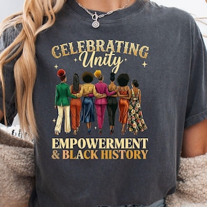May include: Dark gray t-shirt with the words "CELEBRATING Unity" and "EMPOWERMENT & BLACK HISTORY" in gold lettering. The graphic features seven women of color with their arms around each other, wearing various outfits.
