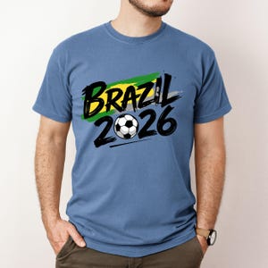 Op de afbeelding: Een blauw t-shirt met de tekst "BRAZIL 2026" in zwart en een voetbalafbeelding. De tekst is over een groen, geel en grijs penseelstrekenontwerp geplaatst. Het shirt heeft een effen blauwe kleur.