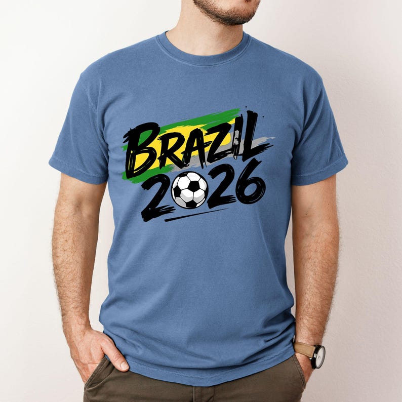 Op de afbeelding: Een blauw t-shirt met de tekst "BRAZIL 2026" in zwart en een voetbalafbeelding. De tekst is over een groen, geel en grijs penseelstrekenontwerp geplaatst. Het shirt heeft een effen blauwe kleur.