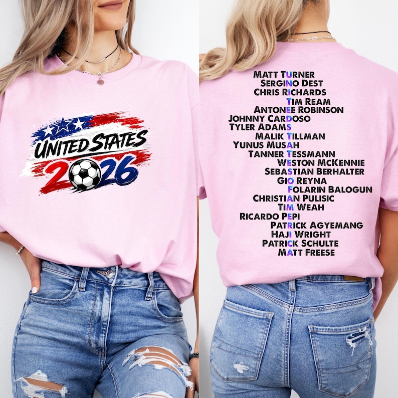 May include: A pale pink t-shirt with the text "UNITED STATES 2026" and a soccer ball design. The back of the shirt has a list of soccer players' names. The shirt is paired with blue jeans.