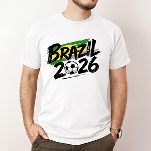 Op de afbeelding: Wit T-shirt met de tekst "BRAZIL 2026" in zwart, en een voetbalafbeelding. Het ontwerp bevat een groene en gele penseelstreek, die de Braziliaanse vlag vertegenwoordigt. Het T-shirt heeft korte mouwen.