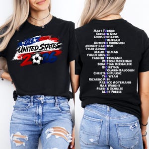 May include: Black t-shirt featuring "UNITED STATES 2026" text and a soccer ball graphic in red, white, and blue. The back of the shirt has a list of names in white. A casual tee for soccer fans.