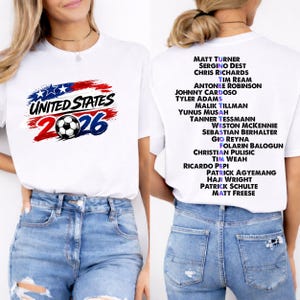 May include: White t-shirt with the text "UNITED STATES 2026" in red, white, and blue, and a soccer ball graphic. The back of the shirt has a list of names. A casual, sporty design.
