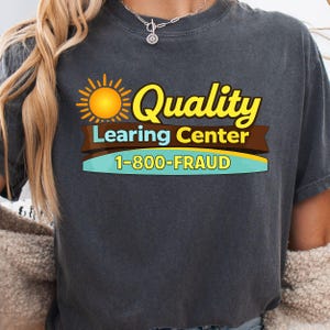 May include: Dark gray t-shirt with a graphic that reads "Quality Learing Center" in yellow and brown, with a sun graphic. Below the text is "1-800-FRAUD" in yellow and blue.