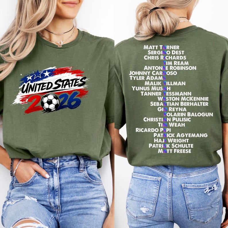 May include: Olive green t-shirt with a graphic featuring the text "UNITED STATES 2026" above a soccer ball. The back of the shirt displays a list of names in white text. A casual, sporty design.