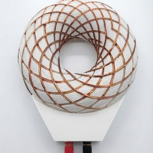 May include: A toroidal coil featuring a white circular core wrapped in a dense, crisscrossing pattern of thin copper wire. The coil is mounted on a white trapezoidal base equipped with one red and one black terminal connector at the bottom edge.