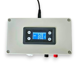 May include: A rectangular light grey electronic control box featuring a central digital display screen with blue text showing OUT 528 050 %. The interface includes four circular buttons labelled F+, F-, D+, and D-. A black power cable extends from the top.