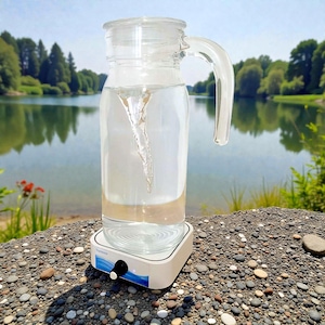 Puede incluir: Una jarra de vidrio transparente con asa y tapa, llena de agua, creando un vórtice visible. La jarra se encuentra sobre una base blanca con un mando de control. El fondo presenta un lago y árboles.