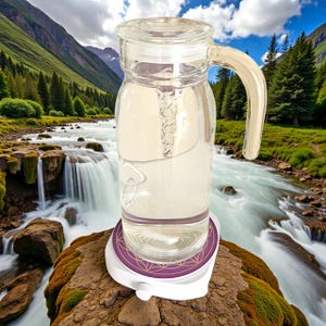 Può includere: Una caraffa d'acqua in vetro trasparente con manico e coperchio, riempita di liquido, poggia su una base bianca e viola. La base presenta un motivo geometrico decorativo. Lo sfondo mostra un fiume e montagne.