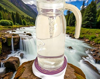 May include: A clear glass water pitcher with a handle and lid, filled with liquid, sits on a white and purple base. The base has a decorative geometric pattern. The background features a river and mountains.