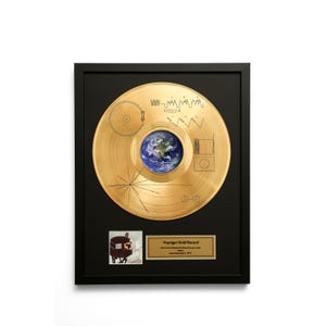 Puede incluir: Disco de oro Voyager enmarcado con marco negro. El disco presenta una imagen de la Tierra en el centro, rodeada de diagramas y símbolos grabados. Una pequeña foto y una placa dorada con texto se encuentran debajo.