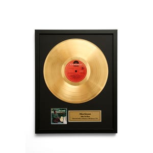 Puede incluir: Disco de oro enmarcado del álbum "Little Girl Blue" de Nina Simone. El disco está centrado en un marco negro, con una pequeña imagen de la portada del álbum y una placa con los detalles del álbum. La etiqueta del disco es roja con texto blanco.