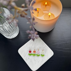 May include: Four pairs of tulip-shaped earrings with red, orange, pink, and white flower beads and green leaf accents. The earrings are displayed on a white marble square, with a lit candle and lavender sprig in the background.
