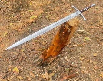 Puede incluir: Una espada larga y plateada con una guarda cruzada y un mango marrón descansa sobre un tronco de madera. La hoja de la espada es afilada y reflectante, y el mango está envuelto en un material oscuro. La escena está ambientada al aire libre, con hojas y agujas de pino en el suelo.