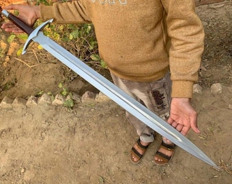 Puede incluir: Una espada larga y plateada con una empuñadura en forma de cruz y un mango marrón oscuro. La hoja de doble filo se estrecha hasta un punto. La espada se sostiene frente a una persona.