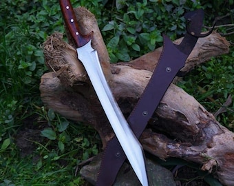 May include: A large, silver-bladed sword with a curved, wooden handle and a brown leather sheath. The handle has a dark wood grain and is accented with small, white rivets. The sheath is dark brown and has a loop for carrying. The sword is resting on a log.