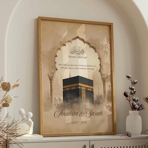 May include: A framed print with a beige background features an illustration of the Kaaba in Mecca. The print includes Arabic calligraphy and the names "Ibrahim & Sarah" with a date. The frame is a light brown color.