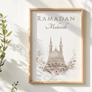 May include: A framed print with the words "RAMADAN Mubarak" in brown script. The image features a mosque with two minarets and a dome, surrounded by a floral wreath in shades of pink, brown, and white. The frame is made of light-colored wood.