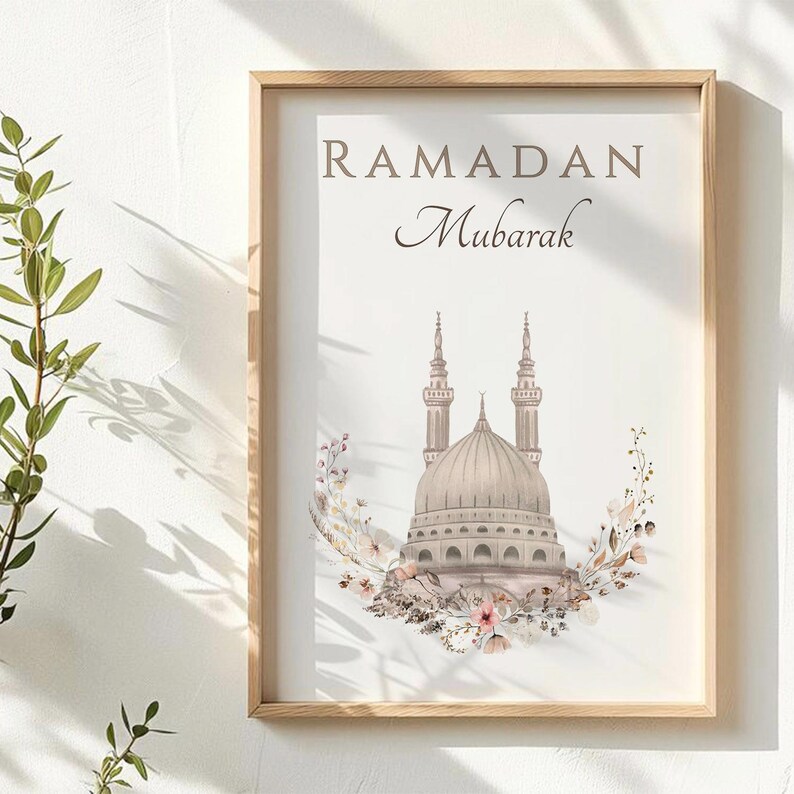 May include: A framed print with the words "RAMADAN Mubarak" in brown script. The image features a mosque with two minarets and a dome, surrounded by a floral wreath in shades of pink, brown, and white. The frame is made of light-colored wood.
