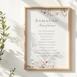 May include: A framed print with the text "RAMADAN Rules of the heart" in a light brown font. The print includes a list of rules and is decorated with floral accents in shades of pink, purple, and brown. The frame is made of light-colored wood.