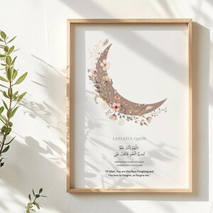 May include: A framed print featuring a crescent moon design with floral accents in shades of brown and pink. The print includes Arabic calligraphy and English text, all within a light wood frame.