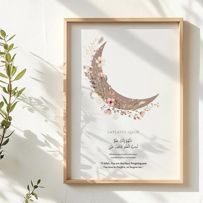 May include: A framed print featuring a crescent moon design with floral accents in shades of brown and pink. The print includes Arabic calligraphy and English text, all within a light wood frame.