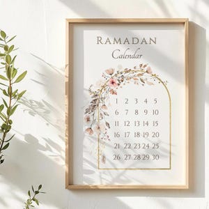 May include: A framed Ramadan calendar with a floral design. The calendar features the word "RAMADAN" above the word "Calendar". The calendar dates are displayed within a gold arch, and the frame is made of light wood.