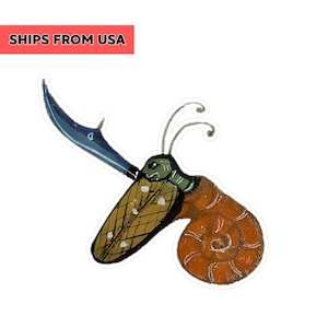 May include: A whimsical sticker featuring a snail with a sword. The snail has a brown shell, a green head, and a yellow body with white dots. The sword is blue. The text "SHIPS FROM USA" is in a red box.