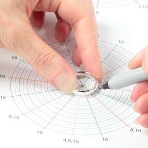 May include: A silver ring is being measured on a ring sizer with a gray marker. The sizer has concentric circles with numbers indicating size.