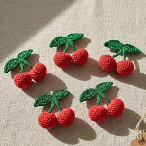 Puede incluir: Cinco cerezas hechas a mano con ganchillo, con hojas verdes y frutos rojos, están dispuestas sobre una superficie de color neutro. Las cerezas están detalladas con una puntada texturizada, mostrando una estética artesanal.