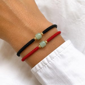 Può includere: Due braccialetti in corda, uno nero e uno rosso, ciascuno caratterizzato da una singola perla quadrata in pietra verde chiaro centrata tra due piccole sfere in metallo dorato. I braccialetti sono realizzati in corda intrecciata liscia.