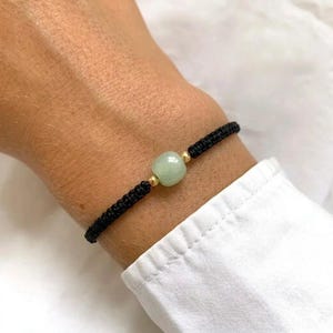 Könnte beinhalten: Ein schwarzes Makramee-Armband mit einer hellgrünen, quadratischen Jadeperle und kleinen goldfarbenen Perlen. Das Armband wird am Handgelenk getragen, mit einem weißen Hemdsärmel im Hintergrund.