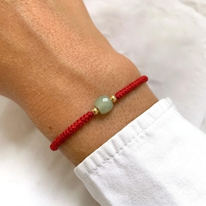 May include: A red braided bracelet with a light green, rounded stone and small gold beads. The bracelet is worn on a wrist, and the background is a white shirt.
