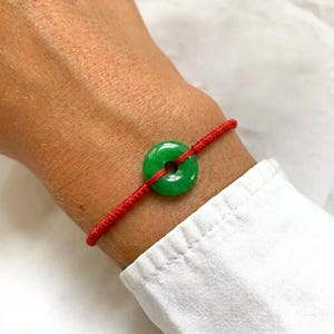 May include: A bracelet featuring a vibrant green, circular jade-like stone bead with a central hole, strung on a thin, bright red cord. The smooth, polished green stone is centred on the wrist, held in place by the simple red string band.
