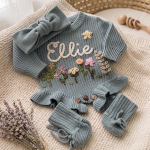Könnte beinhalten: Ein langärmeliger, staubblauer Strickstrampler mit Rüschensaum, drei Holzknöpfen, gestickten Blumendetails und dem Namen Ellie in weißer Schreibschrift. Das Set enthält ein passendes Stirnband mit Knoten und ein Paar Strickschühchen.