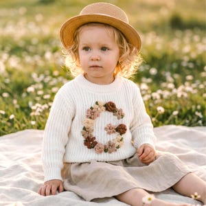 May include: A cream-coloured long-sleeve knit jumper featuring a large letter G formed by embroidered fabric flowers in shades of brown, beige, and cream, paired with a tan straw hat and a light beige skirt.