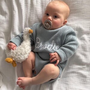 May include: A baby in a light blue knitted romper with the name "Brodie" embroidered in white. The baby holds a white plush goose toy with yellow details and a dummy. The baby is lying on a white textured blanket.
